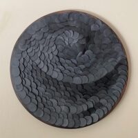The 'New Moon' wall sculpture showcases overlapping dark gray, coin-like discs arranged in a swirling, wave-like pattern against a light wall.