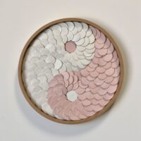 The New Moon wall art features a yin-yang symbol crafted from overlapping white and pink ceramic discs, all set within a circular wooden frame.